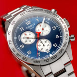The Tissot T1316171104200 PRS 516 Chronograph Men Watch by Tissot Watches features a stainless steel build, blue dial, three subdials, and a tachymeter bezel, shown close-up against a vivid red background.